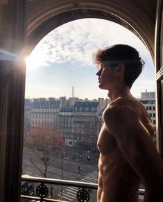 a shirtless man standing in front of an open window looking at the street below