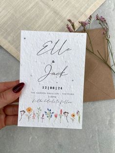 a hand holding up a wedding card with flowers on it and an envelope in the background