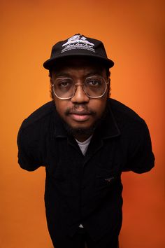 a man with glasses and a hat on posing for a photo in front of an orange background