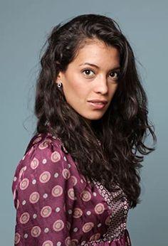 a woman with long dark hair wearing a purple dress looking at the camera while standing in front of a gray background