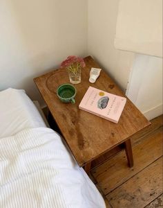 a small wooden table with a book and flower vase on it next to a bed