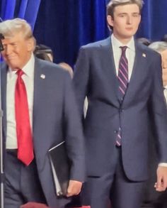 two men in suits and ties walking next to each other with microphones behind them