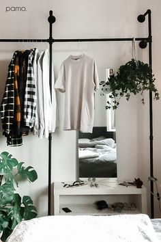 clothes are hanging on a rail in front of a mirror and bed with a plant next to it