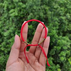 a hand holding a red string bracelet with beaded beads in front of green trees