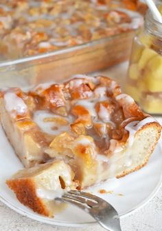 a piece of bread pudding on a plate with a fork next to it and a bowl of fruit in the background