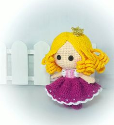 a crocheted doll sitting in front of a white fence with a pink dress