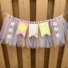 a pink and yellow one year banner hanging from a wooden fence with flowers on it