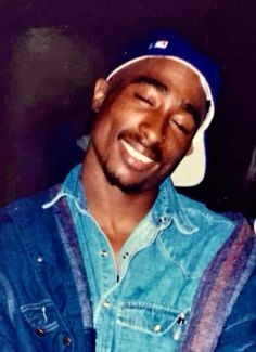 a smiling man wearing a blue jean shirt and hat with his eyes closed while holding a cell phone