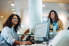 two women sitting at a desk in an office talking to each other while looking at a computer screen