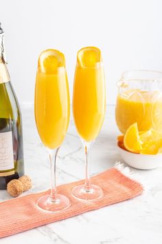 two champagne flutes are sitting on a towel next to a bottle of wine and a bowl of oranges