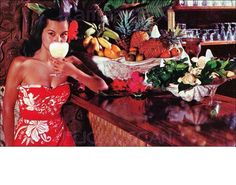 a woman in a red dress standing at a bar with fruit and flowers on it