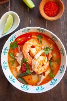 a bowl of shrimp and vegetable soup on a wooden table next to some limes