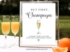 two glasses of champagne and an orange on a table with a sign that says but first, champagne please take a glass for our champagne toast