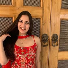 a woman with long brown hair wearing a red dress and smiling at the camera while standing in front of a wooden door