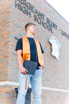 a young man standing in front of a high school building holding a folder and wearing an orange safety vest