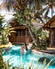 two women are in the pool near some huts and palm trees, one is holding her back