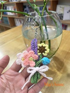 a hand holding a crocheted flower keychain in front of a glass vase