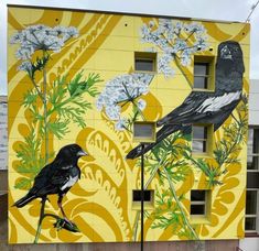 two birds painted on the side of a yellow building with white flowers and green leaves