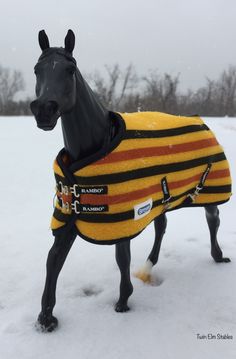 a black horse wearing a yellow and red sweater in the snow with trees in the background