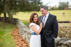 a man and woman standing next to each other in front of a stone wall with trees