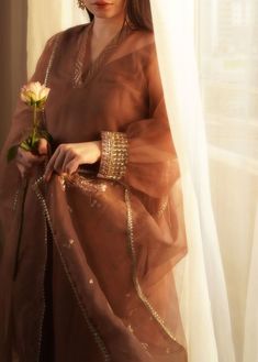 a woman is standing in front of a window with a rose on her hand and wearing a brown dress