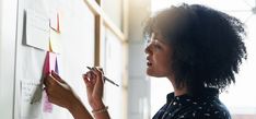 a woman writing on a whiteboard with marker and pen in her hand while another person writes notes on the board