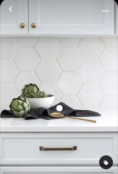 two artichokes in a white bowl on a kitchen counter with gold spoons