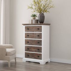 a white dresser with drawers and vase on top in front of a chair next to a window