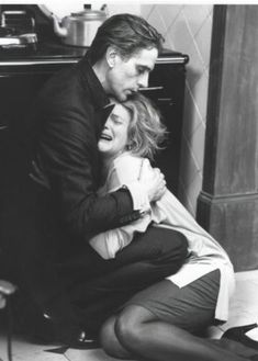 black and white photograph of two people sitting on the floor hugging each other in front of a stove