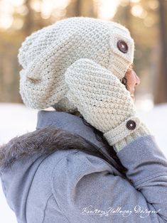 a woman wearing a knitted hat and mittens in the snow with trees behind her