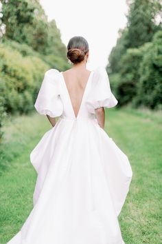 a woman in a white wedding dress walking through the grass with her back to the camera