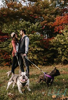 a man and woman walking two dogs on a leash in the grass with trees behind them