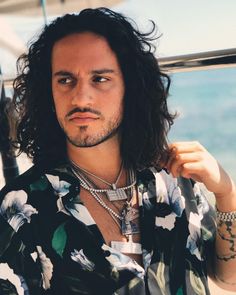 a man with long hair wearing a floral shirt and silver necklace standing in front of the ocean