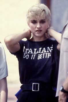 a woman with blonde hair wearing a black t - shirt and silver hoop earrings, standing in front of a group of people