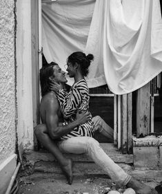 two people sitting on the ground in front of a building and one is kissing the other