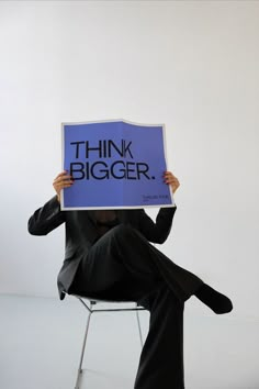 a person sitting in a chair holding up a sign that says think bigger on it