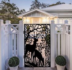 an iron gate with two deers in front of a white house at night, surrounded by potted plants and trees