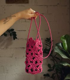 a hand holding a pink crocheted bag in front of a white brick wall