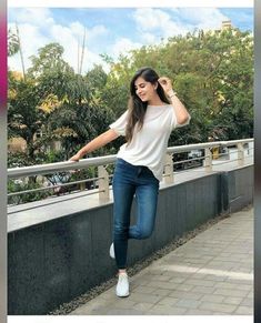 a woman in white shirt and jeans standing on sidewalk next to railing with trees behind her