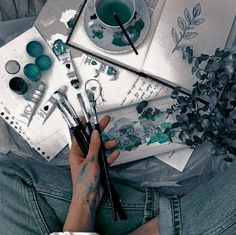 a person with their hands on paintbrushes next to some flowers and other art supplies