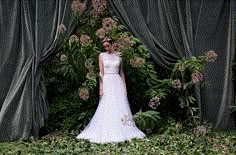 a woman in a wedding dress standing next to some flowers and greenery with curtains behind her