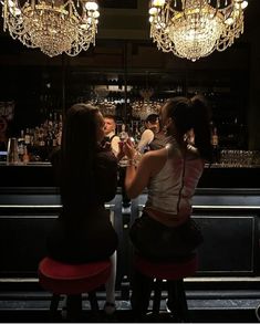 two women sitting at a bar talking to each other with chandeliers hanging from the ceiling