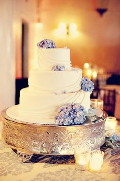 a white wedding cake with blue flowers on the top is sitting on a silver platter