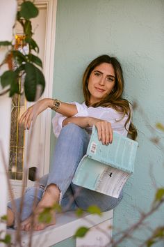 a woman sitting on a window sill holding a blue purse and looking at the camera