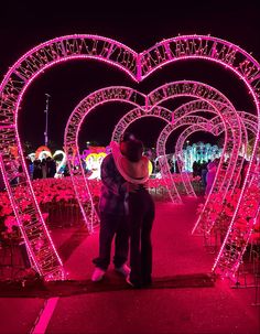 two people standing next to each other in front of pink lights at night with heart shaped archways