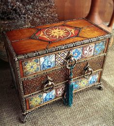 an ornately decorated wooden box with tassels on the lid and handles, sitting next to a potted plant