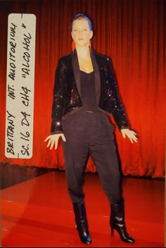 a mannequin is dressed in black and stands on a red carpet with an orange curtain behind it