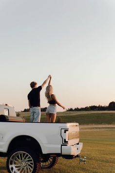 two people standing on the back of a white pickup truck in a grassy field at sunset