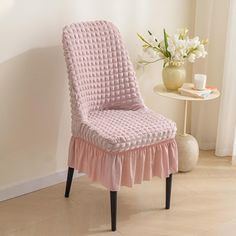 a pink chair sitting in front of a window next to a vase with flowers on it