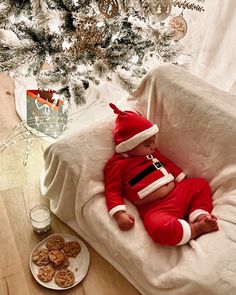 a baby in a santa suit laying on a couch next to a christmas tree